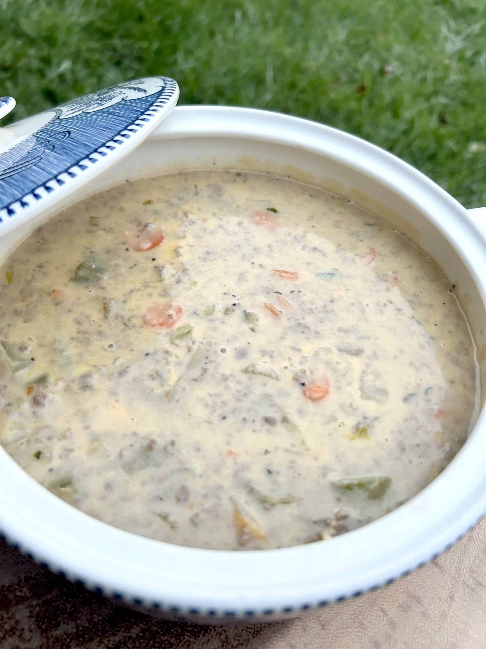 cheeseburger-soup-in-a-small-white-crock-in-the-yard-with-the-lid-to-the-side-