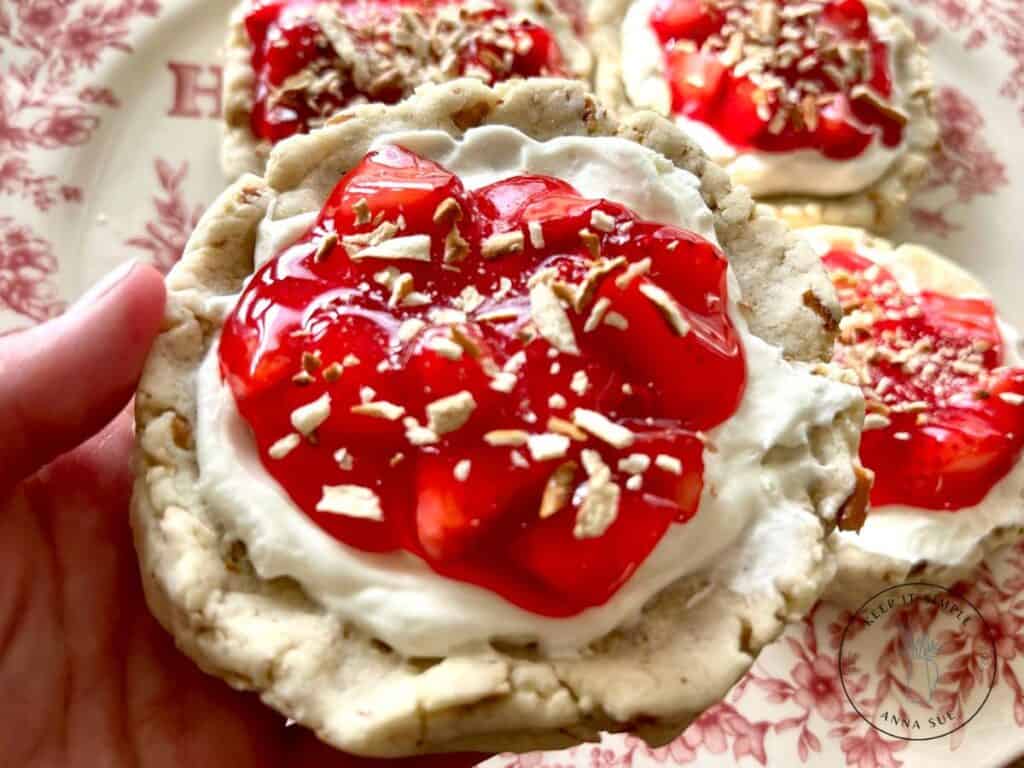 strawberry-pretzel-pie-cookies-with-a-cream-cheese-filling-and-topped-with-a-strawberry-glaze-on-a-beautiful-red-plate-topped-with-pretzel-pieces-with-a-woman-holding-one-cookie.jpg